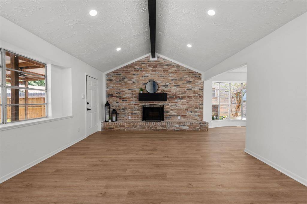 Spacious living room with a vaulted ceiling, featuring a striking brick fireplace wall. The room is brightened by large windows and has modern recessed lighting. The wood like LVT floors add warmth, while the dark beam adds character.