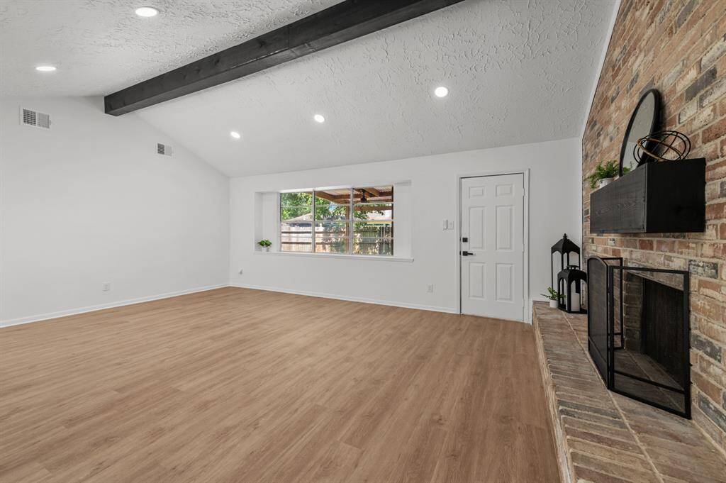 This spacious living room features a vaulted ceiling with a dark wood beam, a cozy brick fireplace, and large windows for natural light. The light-colored wood flooring adds warmth and elegance, creating an inviting atmosphere.
