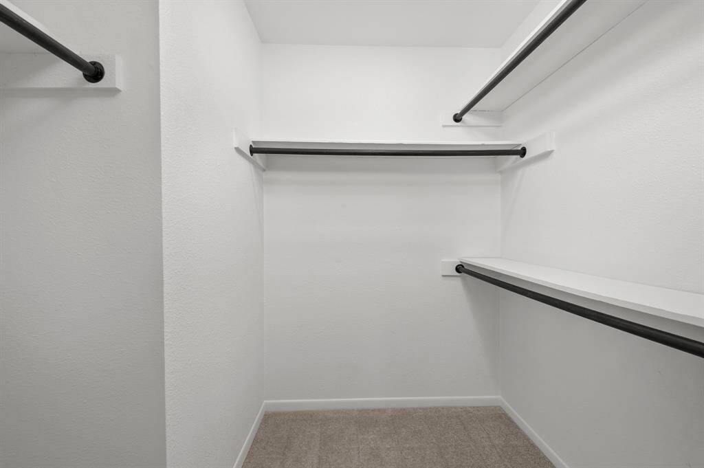 This is a spacious primary bedroom walk-in closet featuring multiple hanging rods on three sides, with clean white walls and a neutral carpeted floor. Perfect for ample clothing storage and organization.