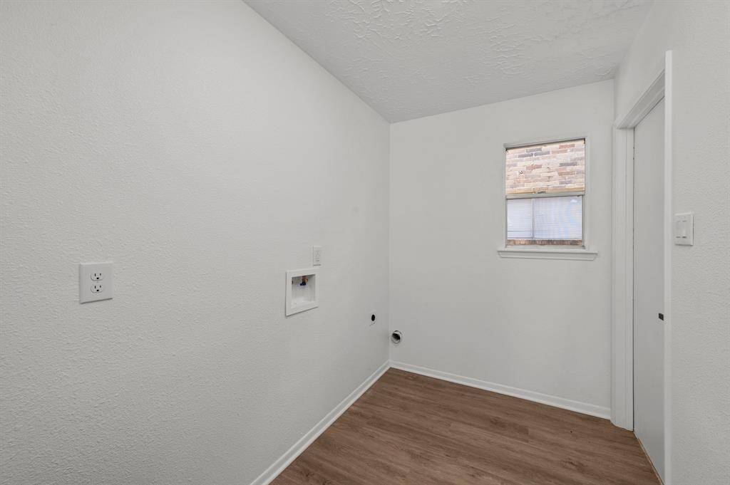 The utility room inside the home, near the kitchen and garage a small, has new white painted walls and wood-like LVT flooring. It has hookups for an electric washer and dryer, a high window for natural light, and a door leading to the garage.