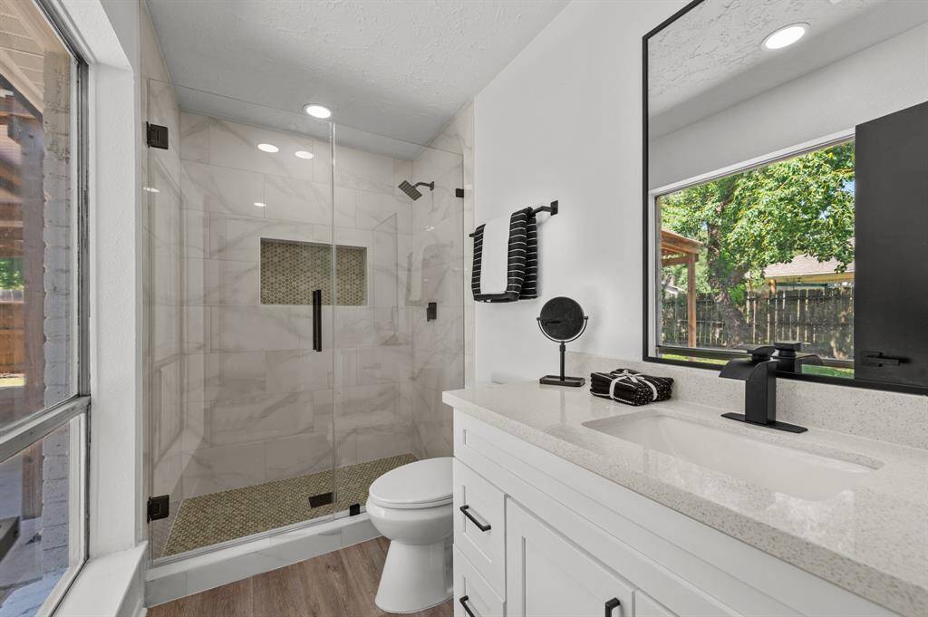 This modern primary bathroom features a spacious glass-enclosed shower with stylish tile work, a sleek vanity with a large mirror, and ample natural light from a window overlooking the backyard.