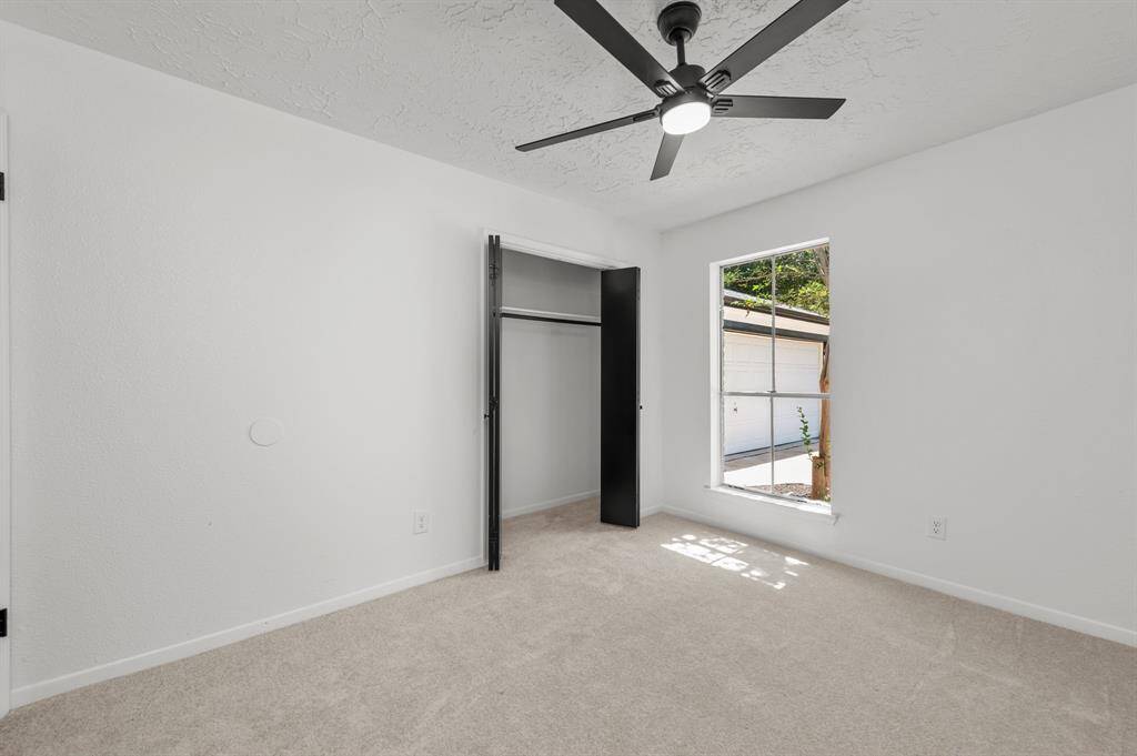 This bedroom features a neutral color scheme with carpet flooring, a ceiling fan, and a large window offering natural light. It includes a closet with black doors, ideal for a bedroom or home office.