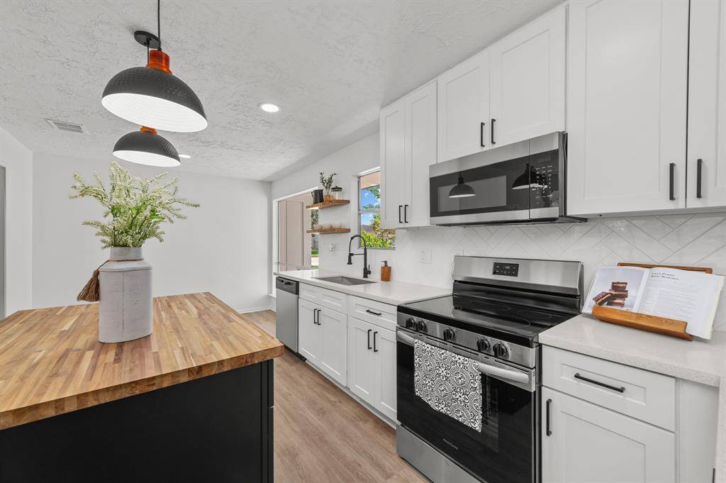 Charming single-story home with a white brick exterior, nestled among mature trees. Features a spacious driveway leading to a two-car garage and a well-maintained front yard, offering both shade and curb appeal.