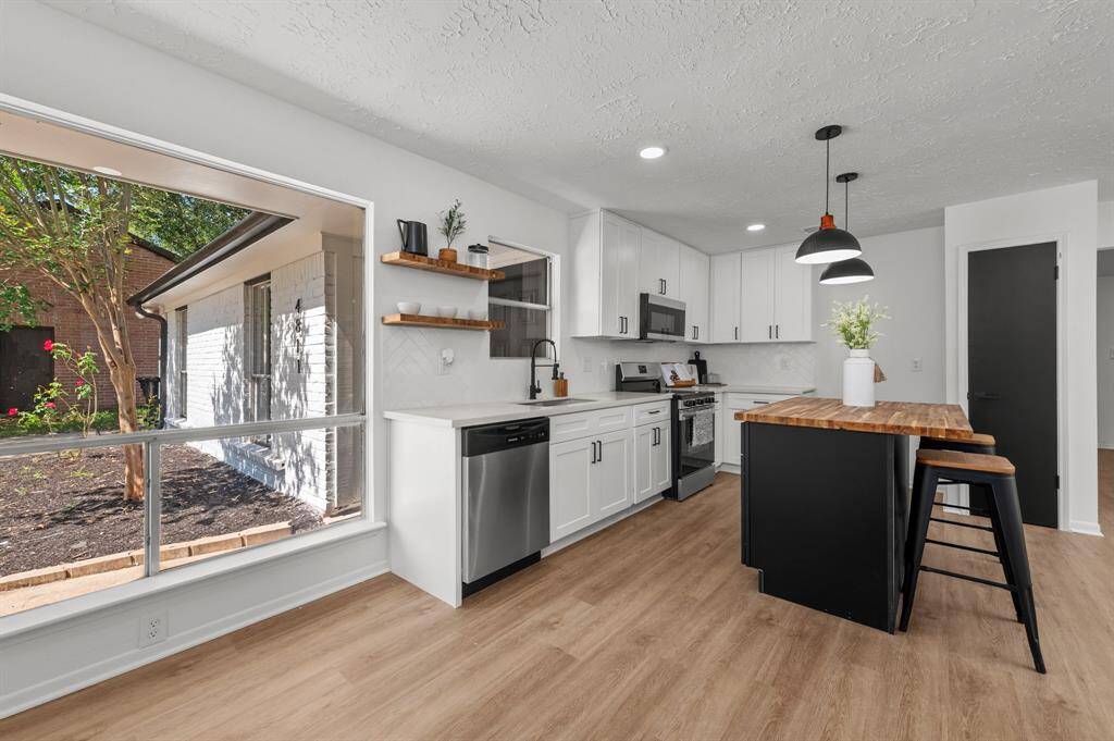 This modern kitchen features sleek white cabinetry, stainless steel appliances, and a stylish butcher block island. Bright and airy, it includes pendant lighting, a herringbone backsplash, and well-placed windows for natural light. Perfect for cooking and entertaining.