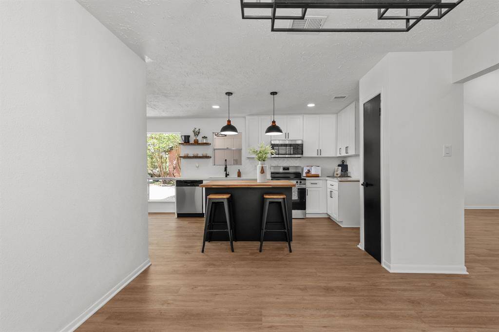 This kitchen is modern and bright, featuring white cabinetry, stainless steel appliances, and a stylish island with bar seating. Large windows offer natural light and a view of the garden, complemented by wood like flooring and pendant lighting for a warm, inviting atmosphere.