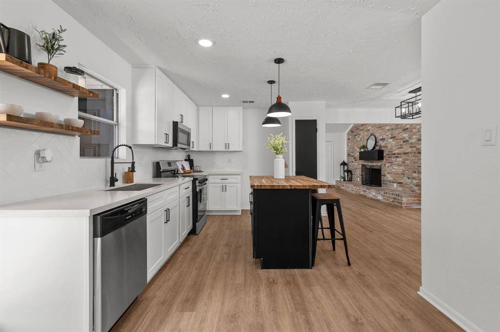 This modern kitchen features sleek white cabinetry, a center island with seating, pendant lighting, and stainless steel appliances. The open layout and wood-style flooring create a warm, inviting atmosphere.