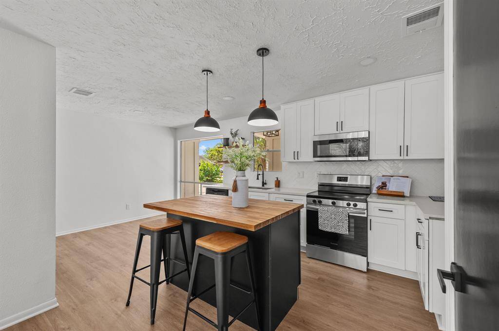 This modern kitchen features sleek white cabinetry, stainless steel appliances, and a stylish island with pendant lighting. The open layout flows into a cozy living area with a huge brick fireplace as the focus of the living room, creating a warm and inviting space.