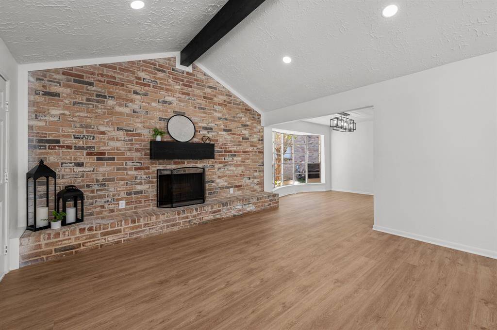 This spacious living area features a stunning brick accent wall with a fireplace, vaulted ceiling with a dark beam, recessed lighting, and beautiful wood like flooring, creating a warm and inviting atmosphere. Perfect for entertaining and for the cozy winter days.