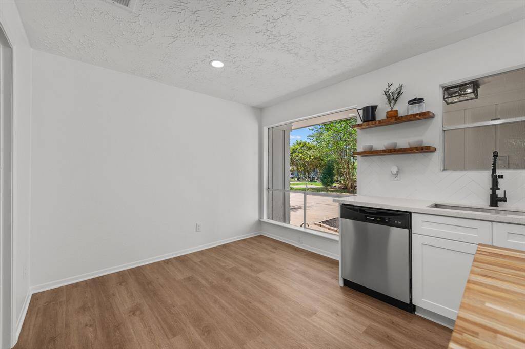 Modern kitchen with white cabinets, stainless steel appliances, and a central island with teak wood butcher block countertop. Features pendant lighting, a herringbone backsplash, and a view of greenery through the window.