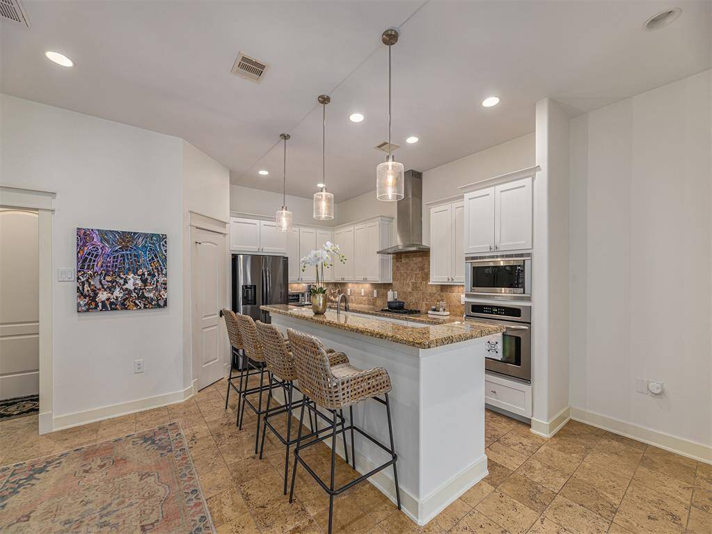 This modern kitchen features a spacious island with seating, granite countertops, and stainless steel appliances. White cabinetry and pendant lighting enhance the bright, open feel, while a colorful artwork adds a touch of character.