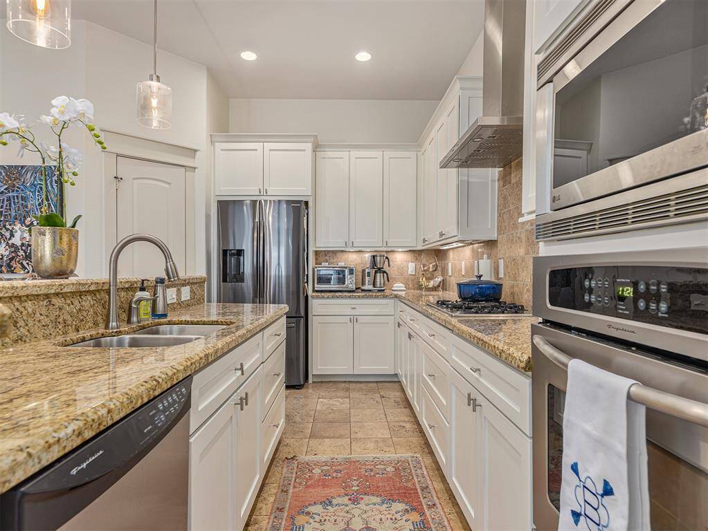 Modern kitchen with granite countertops, stainless steel appliances, and white cabinetry. Features a double sink, gas stove, built-in oven, and microwave. Bright lighting and elegant pendant lights enhance the space.