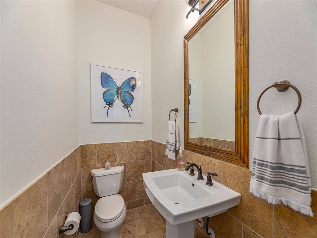 This is a cozy half-bathroom with a pedestal sink, a toilet, and a large mirror. It features a decorative butterfly artwork and warm-toned tile halfway up the walls, giving it a charming and inviting feel.