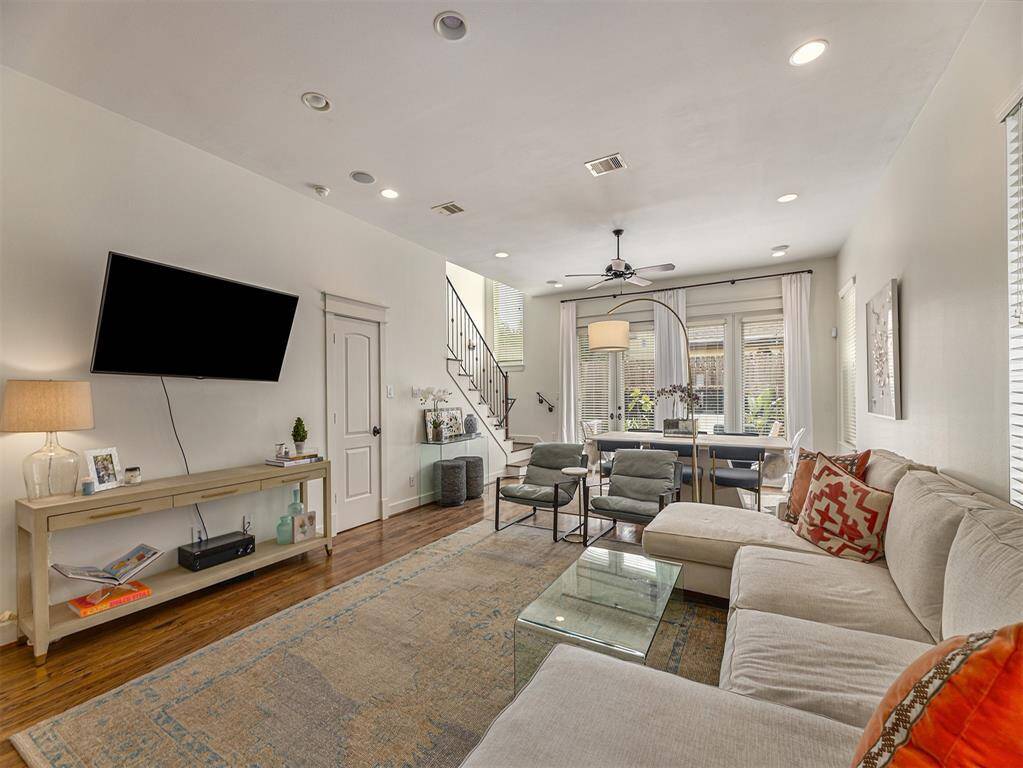 Bright and spacious living room featuring a large sectional sofa, wall-mounted TV, and hardwood floors. The room flows into a dining area with ample natural light from large windows, complemented by modern lighting and cozy decor.