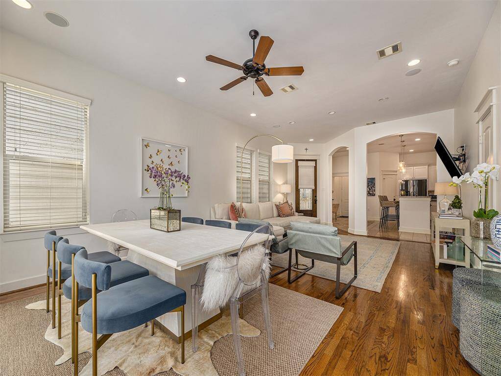 This photo showcases a spacious, open-plan living and dining area with hardwood floors and neutral walls. It features a large dining table with blue chairs, a cozy sofa, a ceiling fan, and ample natural light from multiple windows. The room flows into a modern kitchen with bar seating, creating an inviting space for entertaining.