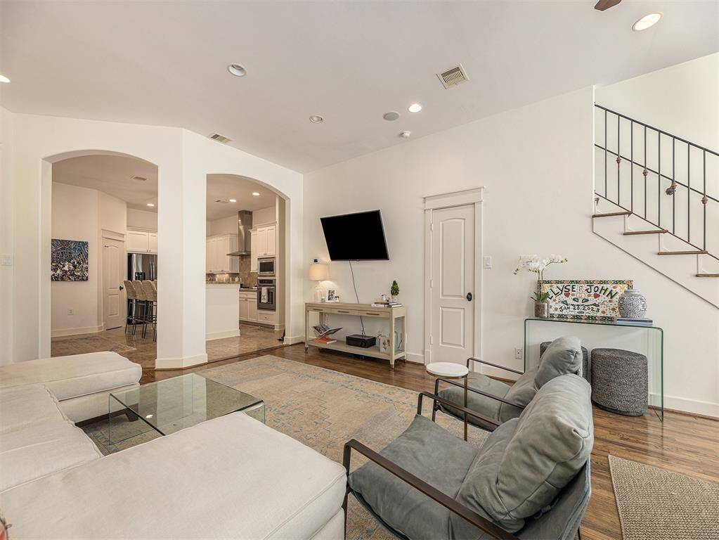 This spacious living room features a cozy seating area with a modern L-shaped sofa, a mounted TV, and stylish decor. An open archway leads to a sleek kitchen with stainless steel appliances. Hardwood floors and a staircase with wrought iron railing add warmth and elegance.