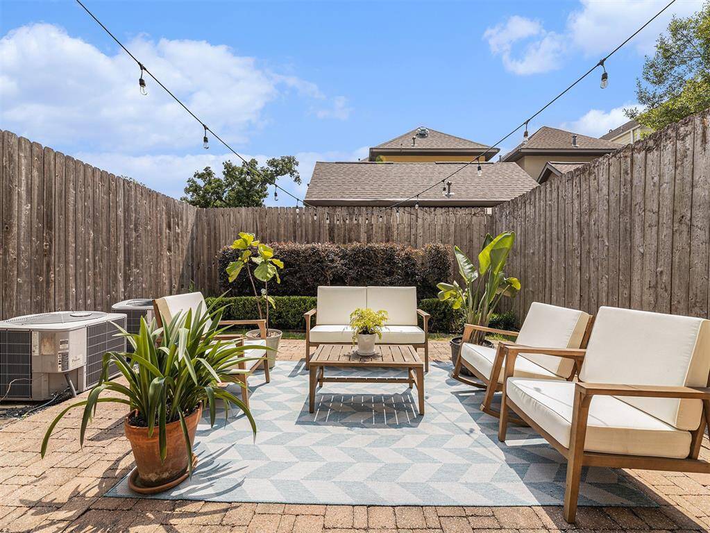 This charming backyard features a cozy seating area with modern outdoor furniture on a patterned rug. Surrounded by a wooden fence, it includes potted plants and string lights, creating a relaxing atmosphere perfect for entertaining.