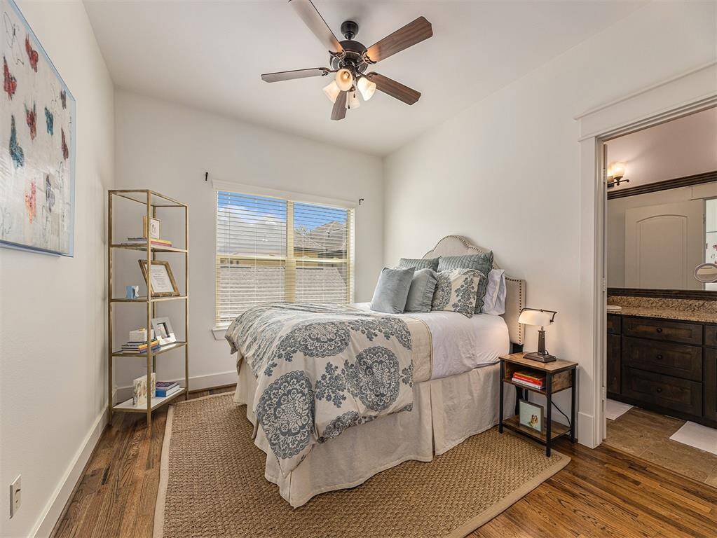 Cozy bedroom with hardwood floors, a ceiling fan, and ample natural light from a large window. Features a comfortable bed, bedside table, and open shelving for storage. En-suite bathroom visible, offering convenience and privacy.