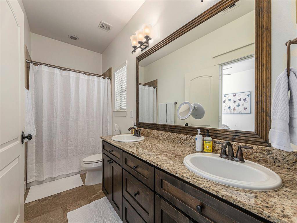 This bathroom features a double vanity with granite countertops, dark wood cabinets, and a large mirror. It includes a shower-tub combo with a white curtain and is well-lit with elegant lighting fixtures. Neutral tones create a warm and inviting atmosphere.