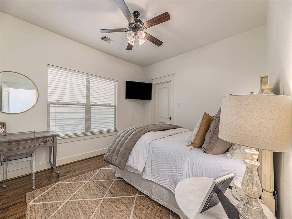 Cozy bedroom with hardwood floors, a ceiling fan, and neutral decor. Features a comfortable bed, a wall-mounted TV, a small desk with a round mirror, and natural light from a large window.