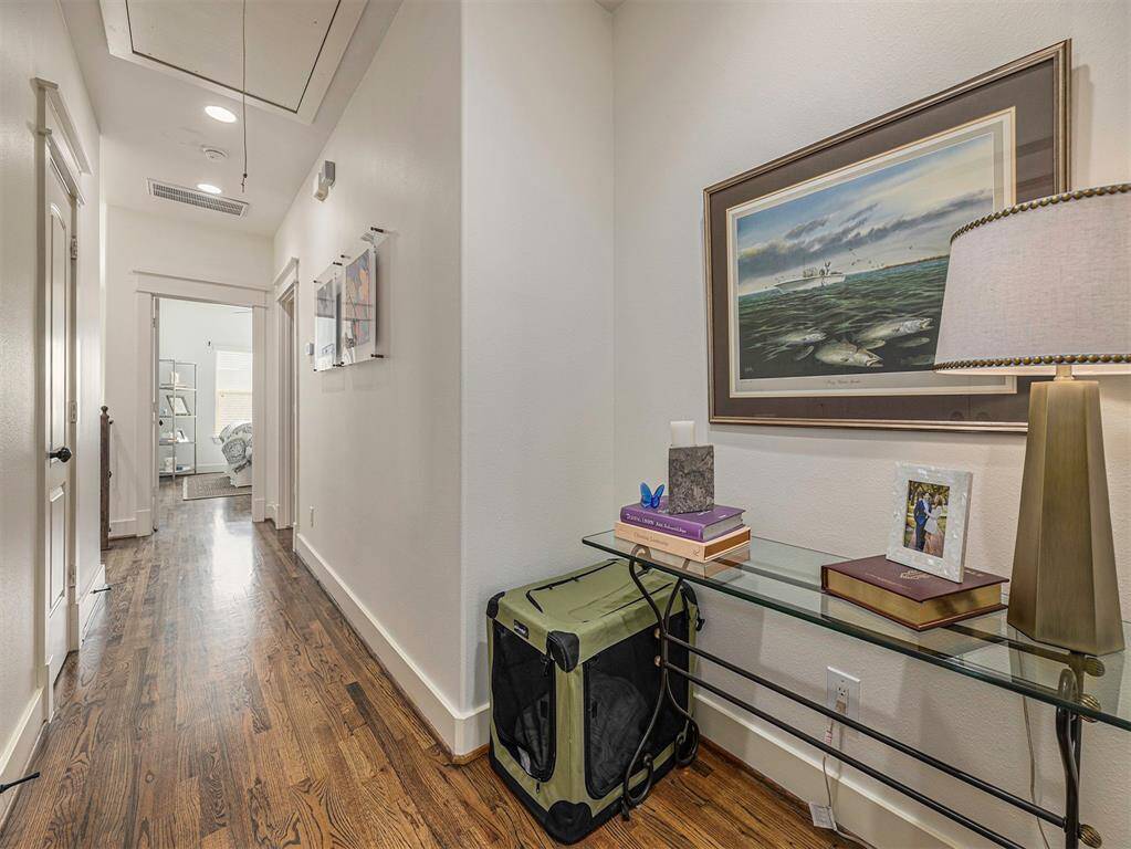 This photo showcases a bright hallway with hardwood floors, leading to a bedroom. The space features a glass side table with decorative items, a wall painting, and recessed lighting, offering a cozy and inviting atmosphere.