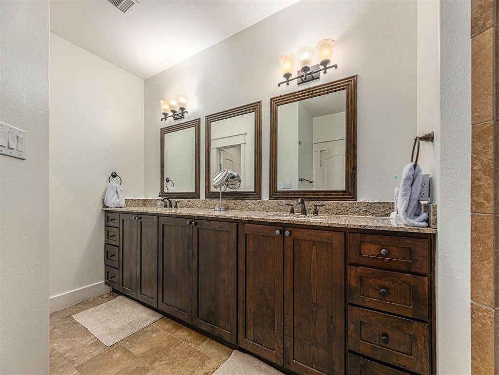 This bathroom features a double vanity with dark wood cabinets, granite countertops, and elegant lighting fixtures above framed mirrors. The neutral tile flooring complements the overall design, providing a warm and inviting atmosphere.