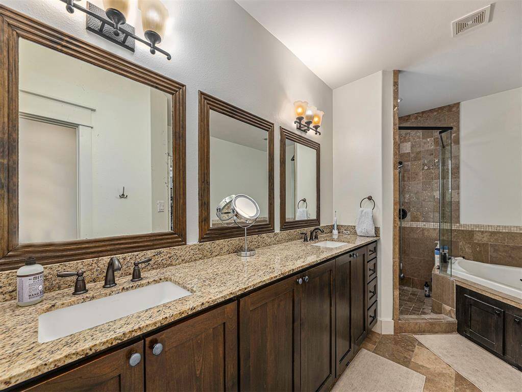 This bathroom features a spacious double vanity with granite countertops and dark wood cabinets. It includes a large mirror, elegant light fixtures, a walk-in glass shower with stone tile, and a cozy bathtub, creating a luxurious and functional space.