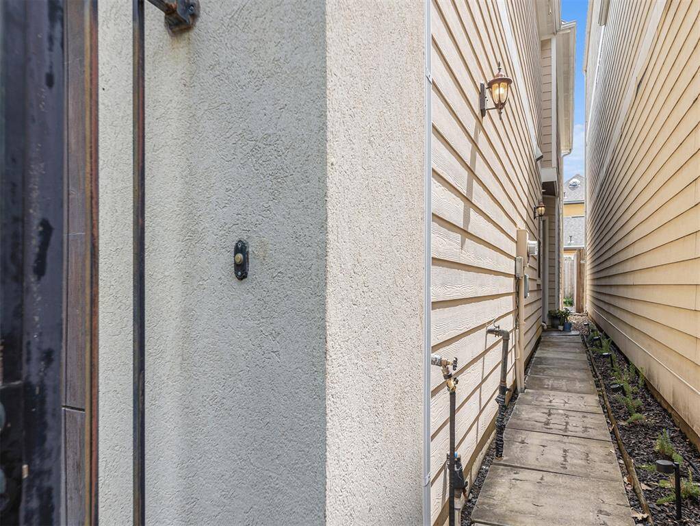 This photo shows a narrow side pathway between two houses, featuring a paved walkway, wall-mounted light fixtures, and a small garden area.