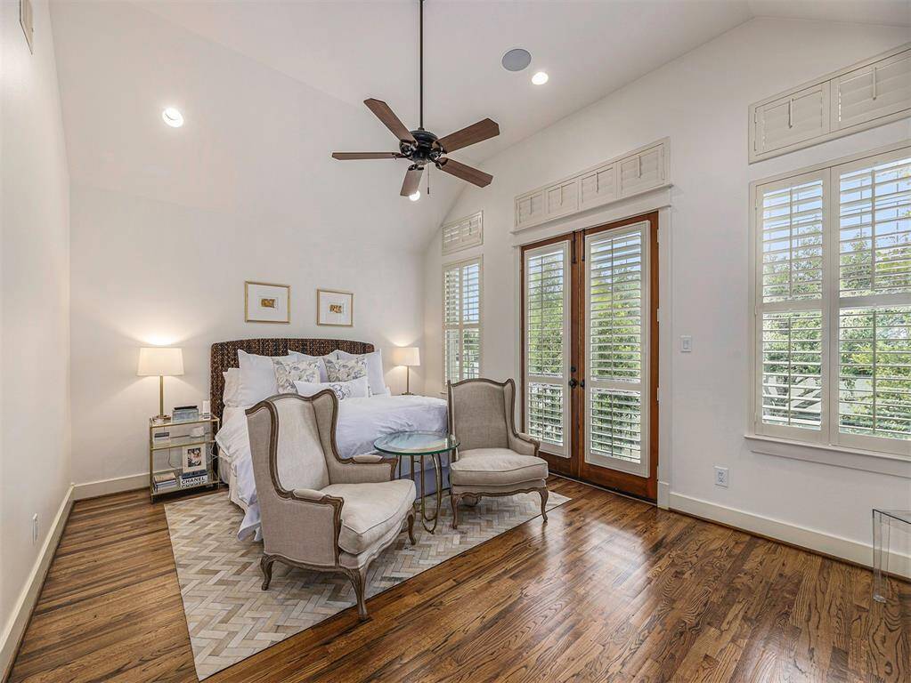 This inviting bedroom features hardwood floors, high ceilings, and ample natural light from large windows with plantation shutters. A ceiling fan complements the cozy setup, which includes a comfortable bed and elegant seating area. French doors lead to a private outdoor space, enhancing the room's appeal.