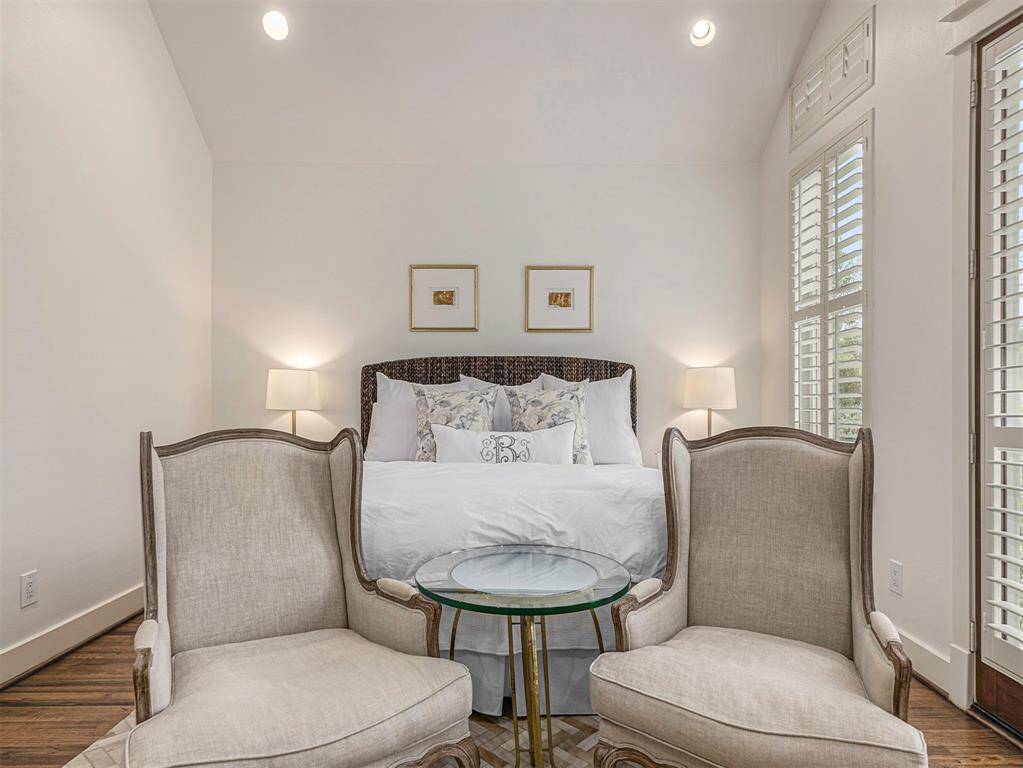 This cozy bedroom features a high ceiling, soft lighting, and neutral tones. It includes a comfortable bed with decorative pillows, two elegant armchairs with a glass table, and large windows with shutters for natural light.
