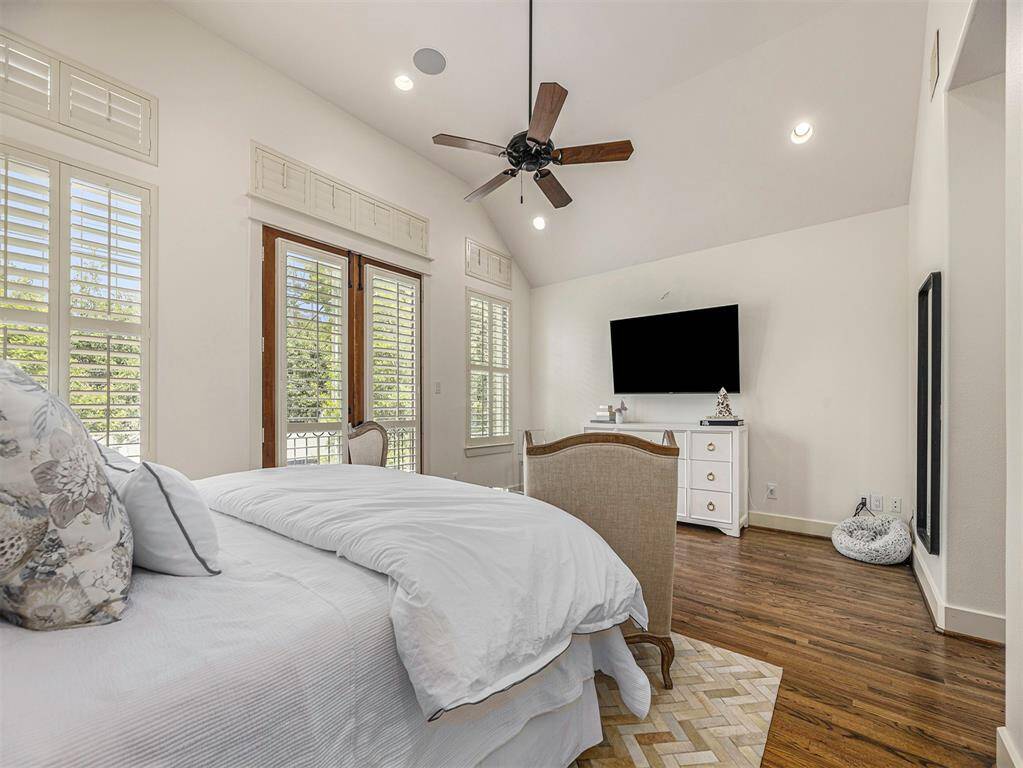 Bright and spacious bedroom with vaulted ceilings, hardwood floors, and large windows with shutters. Features a ceiling fan, wall-mounted TV, and elegant furnishings, creating a cozy and inviting atmosphere.