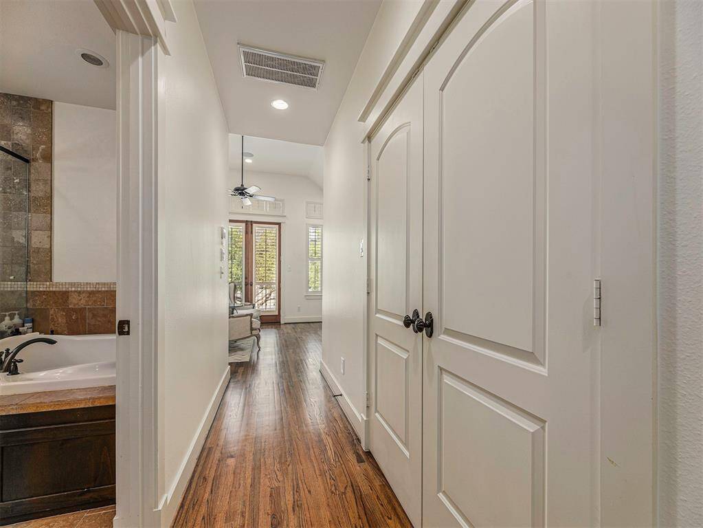 This photo shows a hallway with hardwood floors, leading to a room with French doors and large windows. On the left, there's a glimpse of a bathroom with a bathtub and tiled walls. The hallway includes a closet with white double doors.