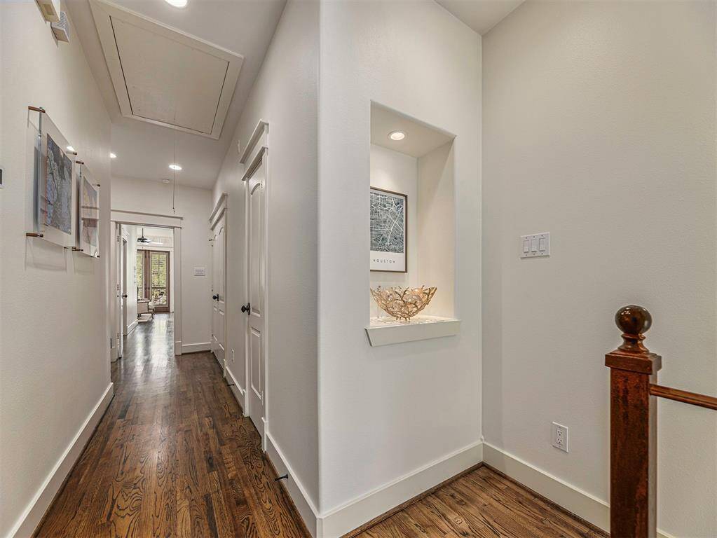 This photo showcases a bright, inviting hallway with polished hardwood floors, recessed lighting, and tasteful wall art. A decorative niche adds character, while the view leads to a well-lit room with large windows, enhancing the home's spacious feel.