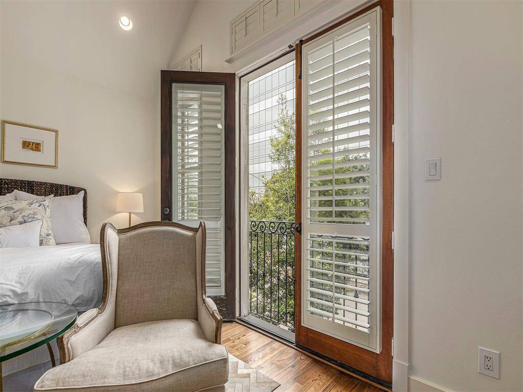 This bright bedroom features elegant French doors with shutters leading to a small balcony, offering a view of greenery. The room is cozy and well-lit, with a comfortable armchair and soft bedding, creating a welcoming and serene atmosphere.