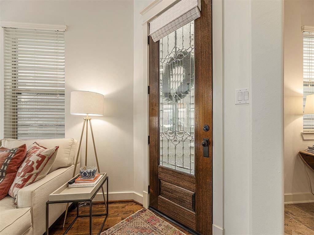 This inviting entryway features a beautifully crafted wooden front door with decorative glass panels. Adjacent is a cozy living area with a comfortable sofa, accent pillows, and a stylish floor lamp. The space is well-lit with natural light from the windows, creating a warm and welcoming atmosphere.