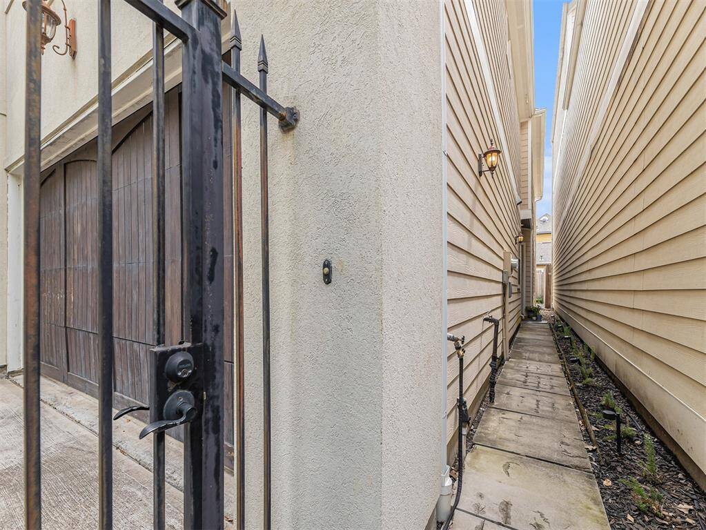 This image shows a narrow side walkway between two homes with beige siding. A black wrought iron gate and a wooden garage door are visible on the left. The path is lined with small plants and has outdoor lighting.