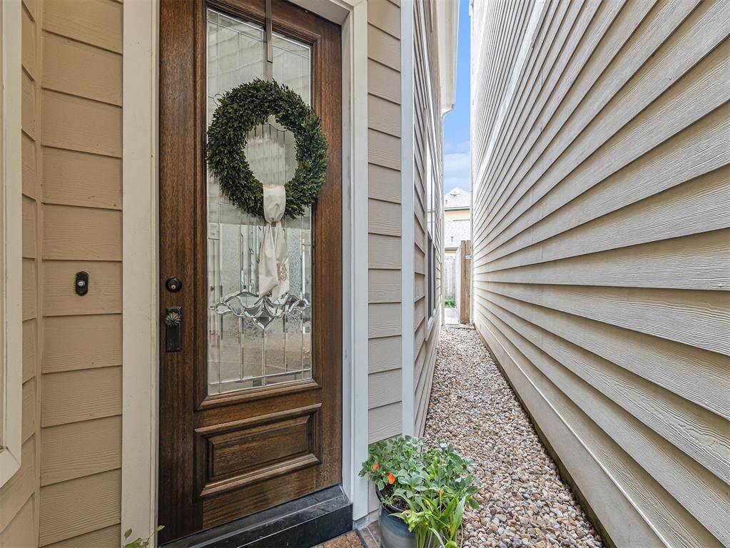 The photo showcases a welcoming front entrance with a wooden door featuring decorative glass and a wreath. The entryway is bordered by light-colored siding, leading to a narrow gravel path with a small planter, suggesting a cozy and low-maintenance exterior space.