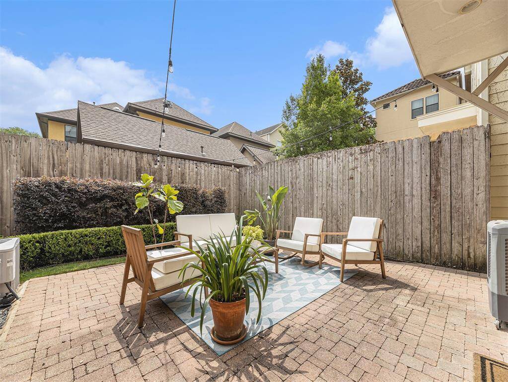 Cozy backyard patio with brick flooring, surrounded by a wooden fence for privacy. Features comfortable outdoor seating, a small rug, and lush plants, perfect for relaxing or entertaining.