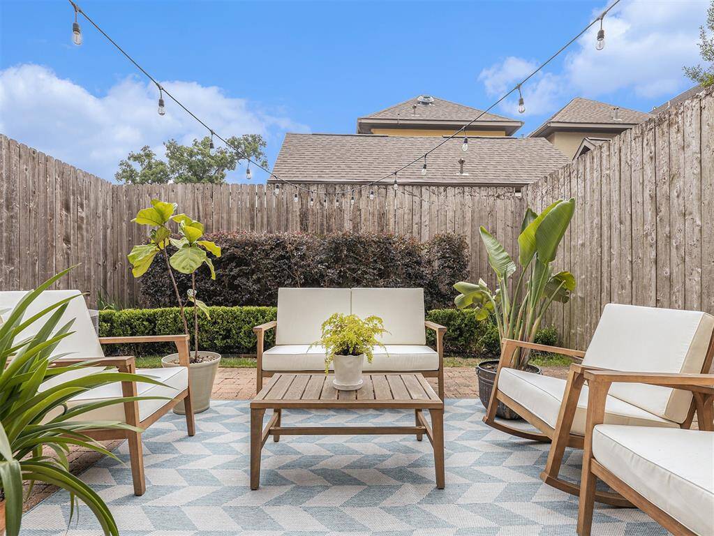 This photo showcases a cozy outdoor patio with comfortable seating, a wooden table, and string lights. The space is surrounded by a wooden fence and lush greenery, creating a private and inviting atmosphere.