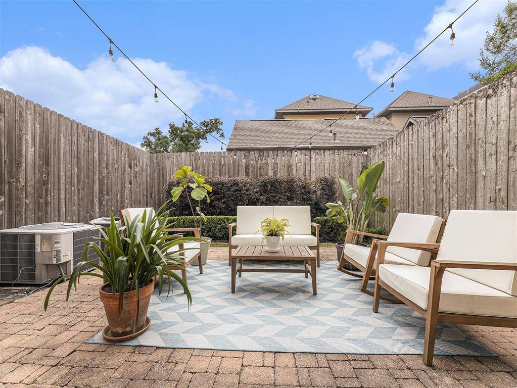 This cozy backyard features a brick patio with outdoor seating and a coffee table on a patterned rug. It’s enclosed by a wooden fence, adorned with string lights, and complemented by potted plants, creating a private and inviting space.