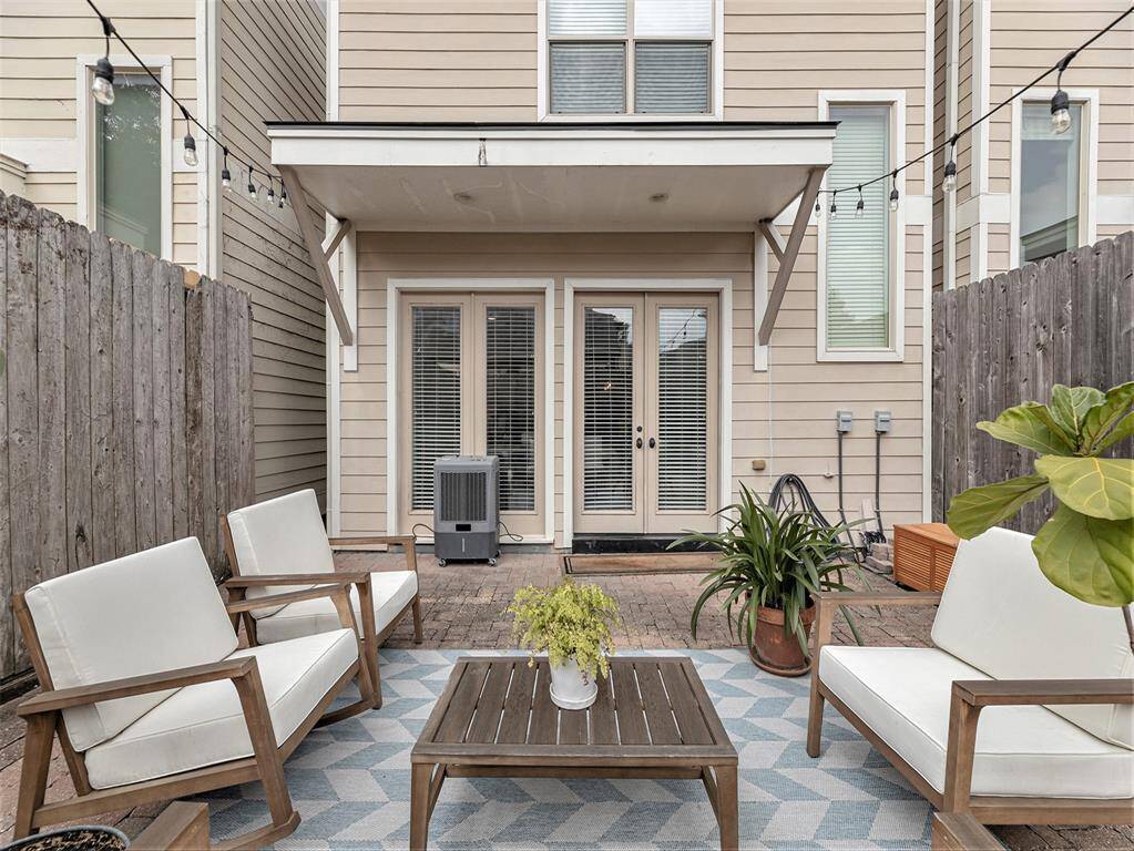 This photo showcases a cozy and inviting backyard patio with a seating area featuring white cushioned chairs and a wooden coffee table. The space is enclosed by wooden fencing, and string lights add a warm, ambient touch. French doors lead into the home, offering a seamless indoor-outdoor flow.