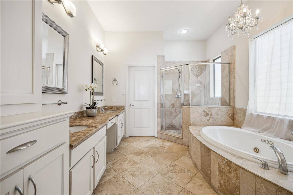 Elegant primary bathroom featuring a large vanity with dual sinks, decorative mirrors, and vanity lights. The space includes a vanity knee space & ample storage. Private water closet. Notice the large jetted tub with a decorative chandelier above for a touch of glamour, and a large window offering natural light. The walk-in shower has a glass door and tile surround. The door ahead opens to a spacious walk-in closet with a dresser and a full-length mirror, completing this luxurious retreat.