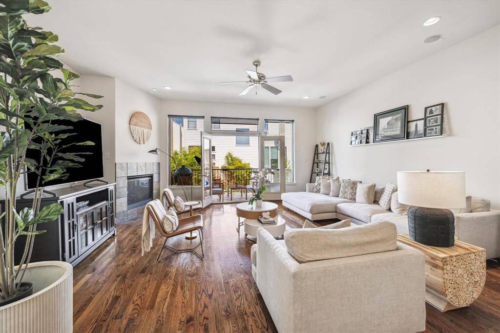 Bright and airy living room with a wall of windows and French doors opening to the large balcony overlooking the backyard. Features include: hardwood floors, a ceiling fan, recessed can lighting, and an art light over the gas log fireplace with a stone surround. Enjoy music through ceiling speakers and tons of natural light from large windows overlooking the lush backyard. The windows come with pull-down shades, while the newly redone patio space boasts a wood-look tile floor. Step out onto the spacious bal