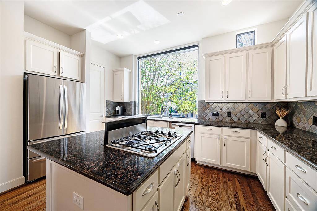 Bright and airy kitchen with a large window overlooking mature trees, an island with barstool seating, and a separate breakfast bar. Features granite countertops, a stone backsplash, ample storage, and under-cabinet lighting, creating a perfect space for cooking and entertaining. Notice the NEW LG refrigerator with two freezer drawers (2024). Door to the left leads to the pantry.