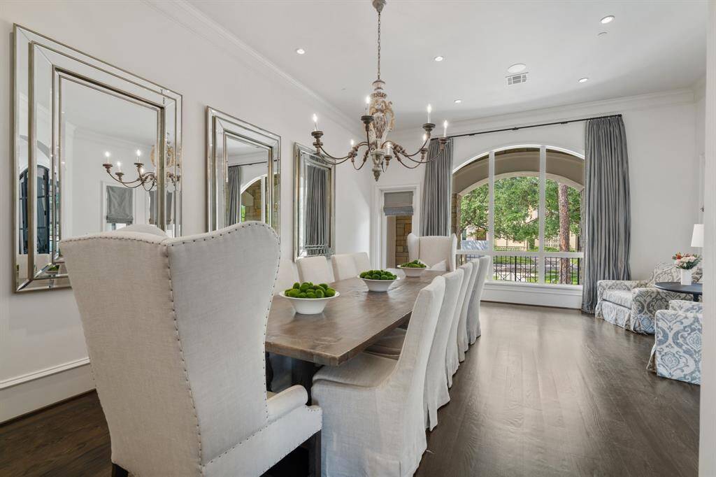 This refined formal dining room exudes timeless sophistication with its rustic wood table, slipcovered seating, and antique-inspired chandelier anchoring the space. A trio of statement mirrors adds depth and elegance, while the grand arched window bathes the room in natural light and frames serene views of the manicured front grounds.