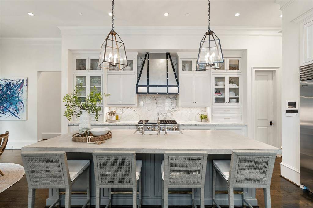 This impeccably designed kitchen pairs classic elegance with modern edge, featuring a bold custom metal range hood, striking marble backsplash, and glass-front cabinetry for refined display. A large center island with seating for four invites casual gatherings beneath statement pendant lighting, creating a space as functional as it is stylish.