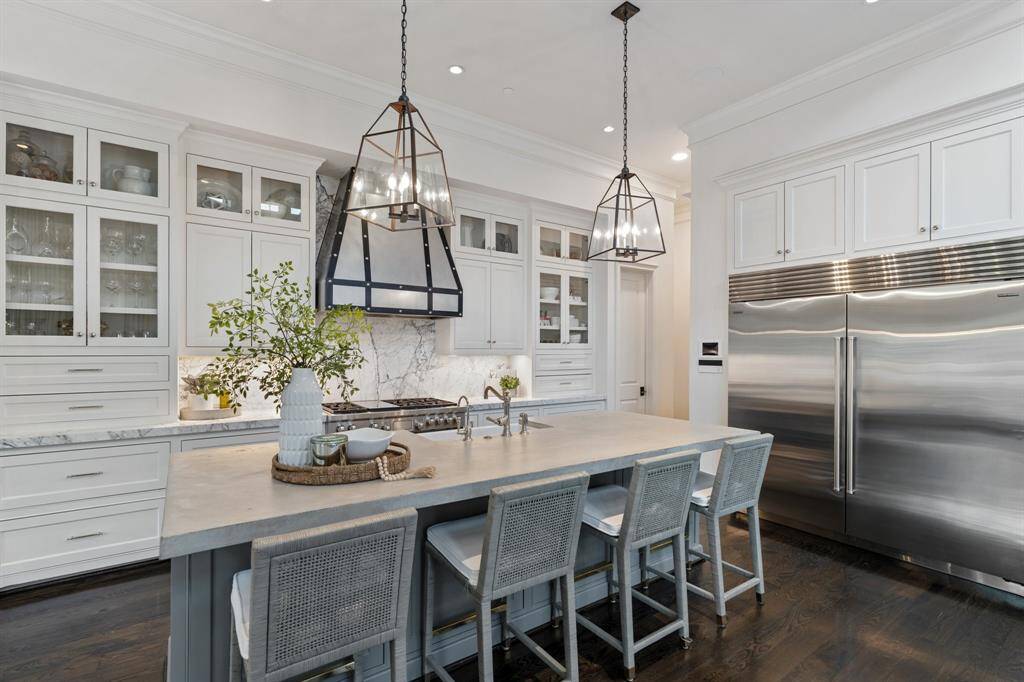 This alternate angle highlights the kitchen’s seamless blend of luxury and functionality, featuring a commercial-grade stainless steel refrigerator, custom glass-front cabinetry, and expansive marble countertops. The oversized island with integrated sink and elegant pendant lighting anchors the space, offering a perfect setting for both meal prep and casual gatherings.