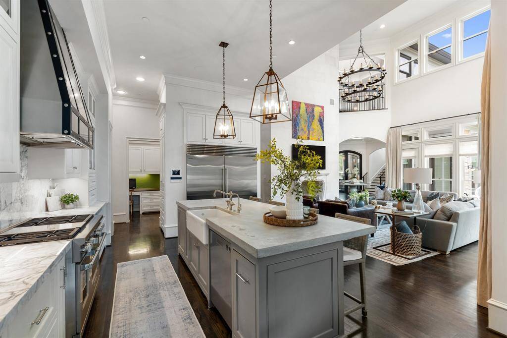 This expansive perspective showcases the seamless flow between the state-of-the-art kitchen and the soaring great room, where layered textures, dramatic ceiling height, and abundant natural light create a captivating ambiance. Designer pendant lighting, a farmhouse sink, and professional-grade appliances enhance the kitchen’s functionality, while sightlines to the cozy living area and colorful artwork add vibrancy and warmth to the heart of the home.