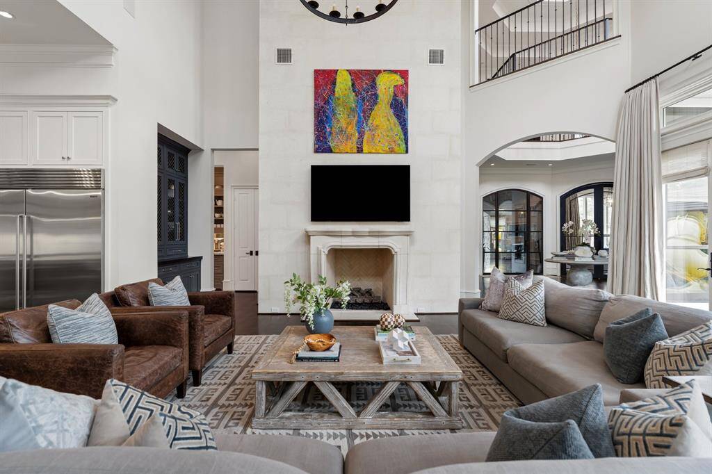 This impeccably styled great room centers around a striking floor-to-ceiling limestone fireplace with a herringbone firebox and mounted TV, creating a focal point of modern luxury. Flanked by tailored furnishings, layered textiles, and captivating architectural views into the gallery-style entry, the space exudes warmth, sophistication, and effortless flow for everyday living and entertaining.