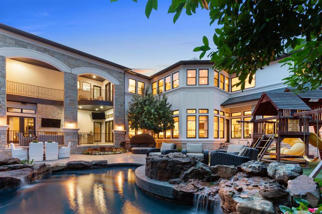 This breathtaking view of the backyard retreat showcases the perfect blend of luxury and leisure, framed by the elegant architecture of the estate’s rear façade. A glistening freeform pool with cascading rock waterfalls anchors the space, surrounded by stylish lounge seating, lush landscaping, and an inviting children’s playset. Illuminated windows and arched stone columns reflect the warm glow of the interior, while the covered patio and balcony offers a serene vantage points for outdoor.