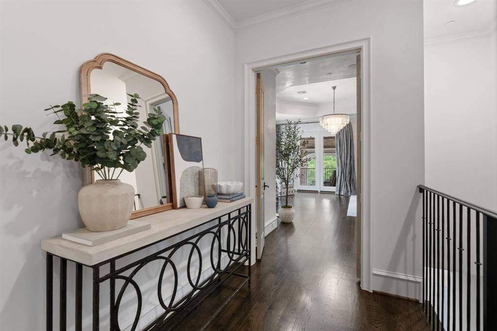 This elegant hallway leading to the primary suite is adorned with a curated console vignette and artful décor, offering a graceful transition framed by rich hardwood floors and a glimpse of the crystal chandelier beyond.